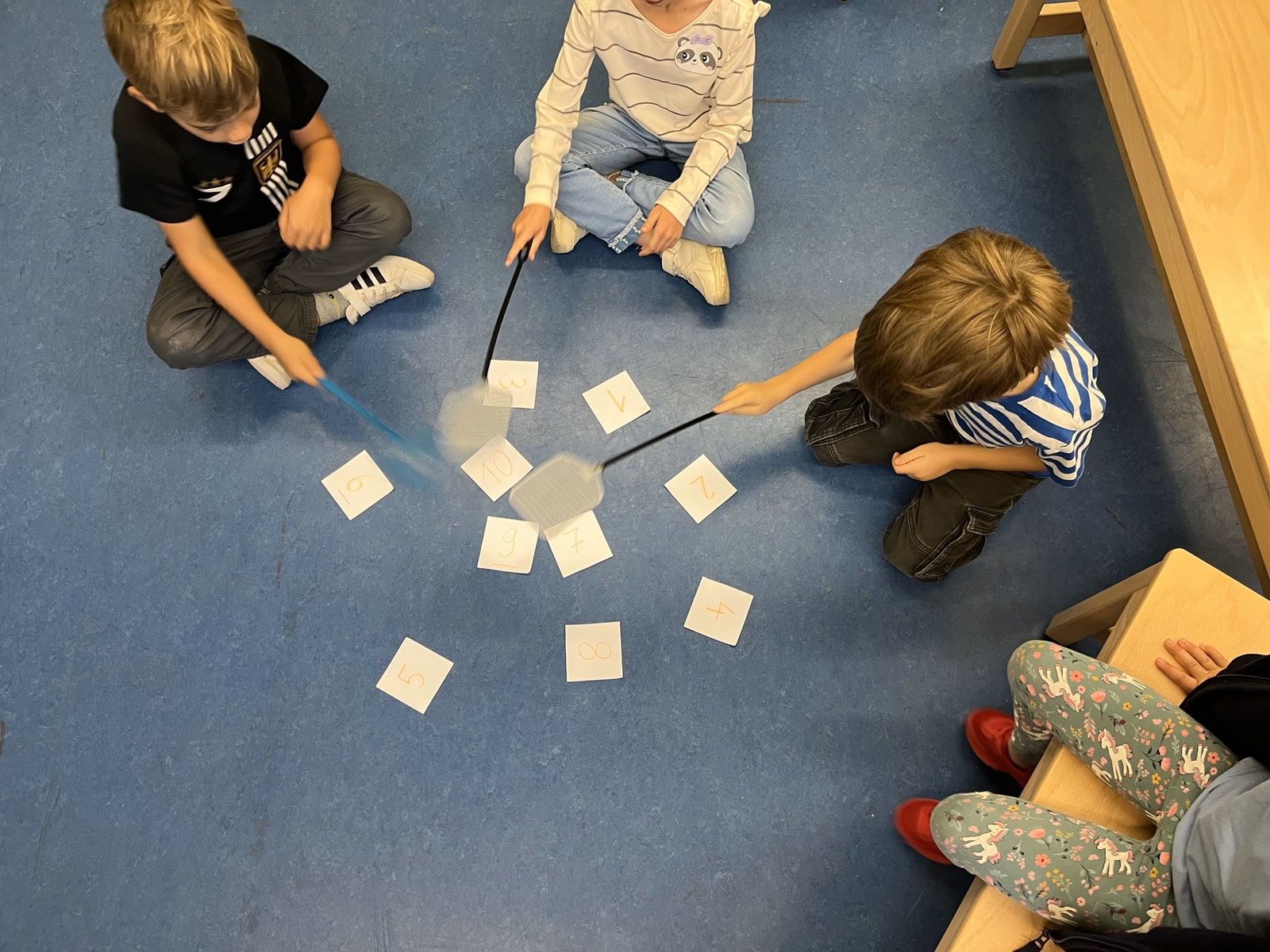 Drei Kinder spielen auf dem Boden mit Fliegenklatschen und Zahlenkarten